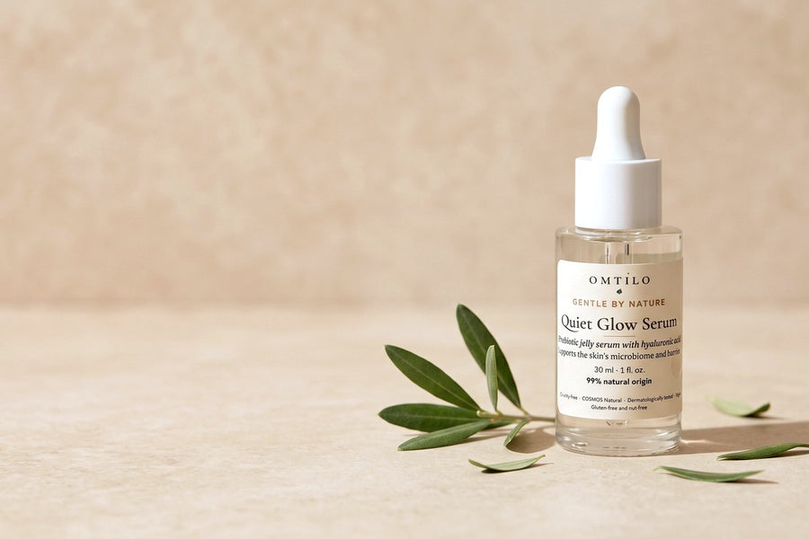 The OMTILO Quiet Glow Serum beside a green olive sprig and scattered leaves